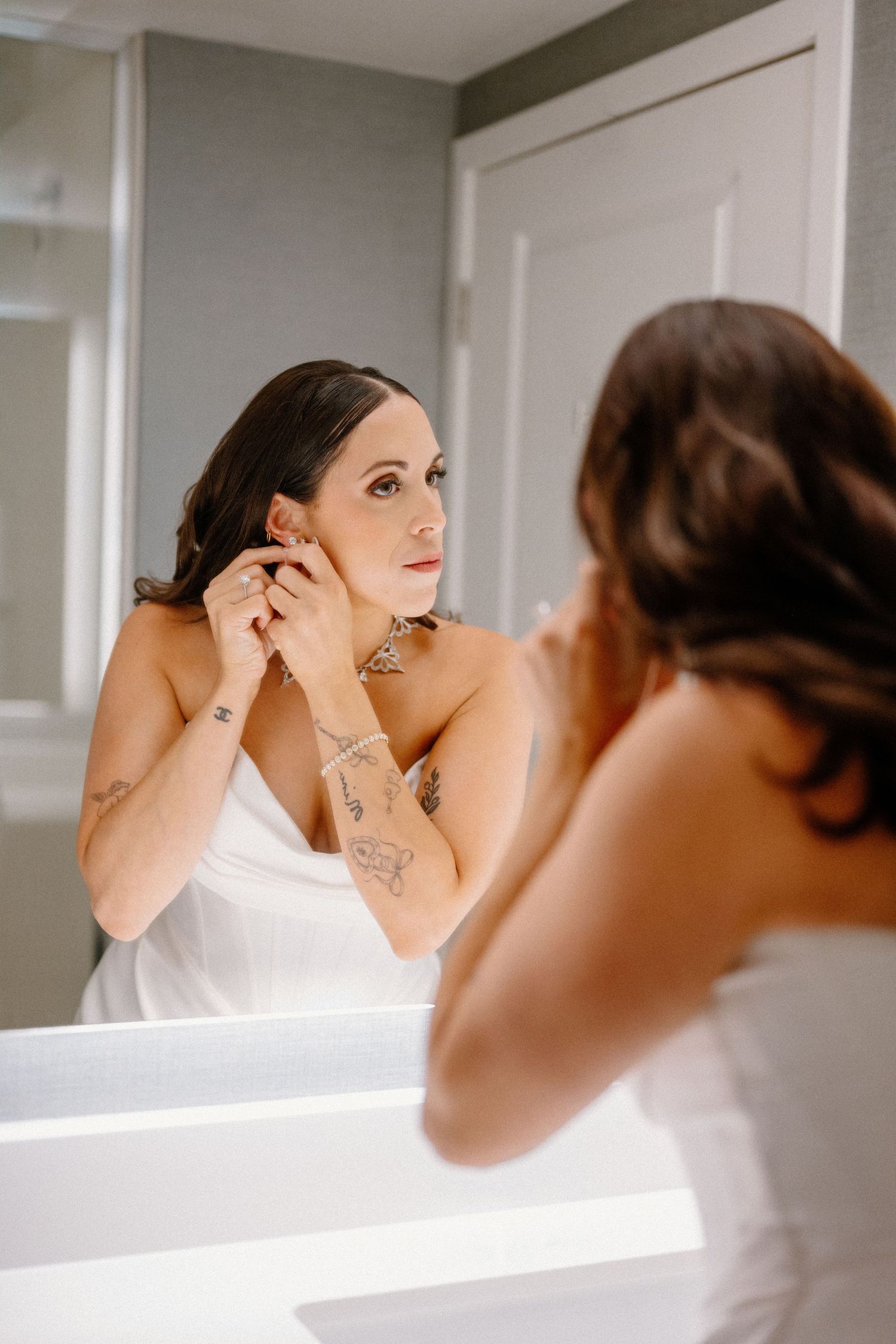 Bride putting on earrings while looking in a lit bathroom mirror, wearing a white strapless dress.