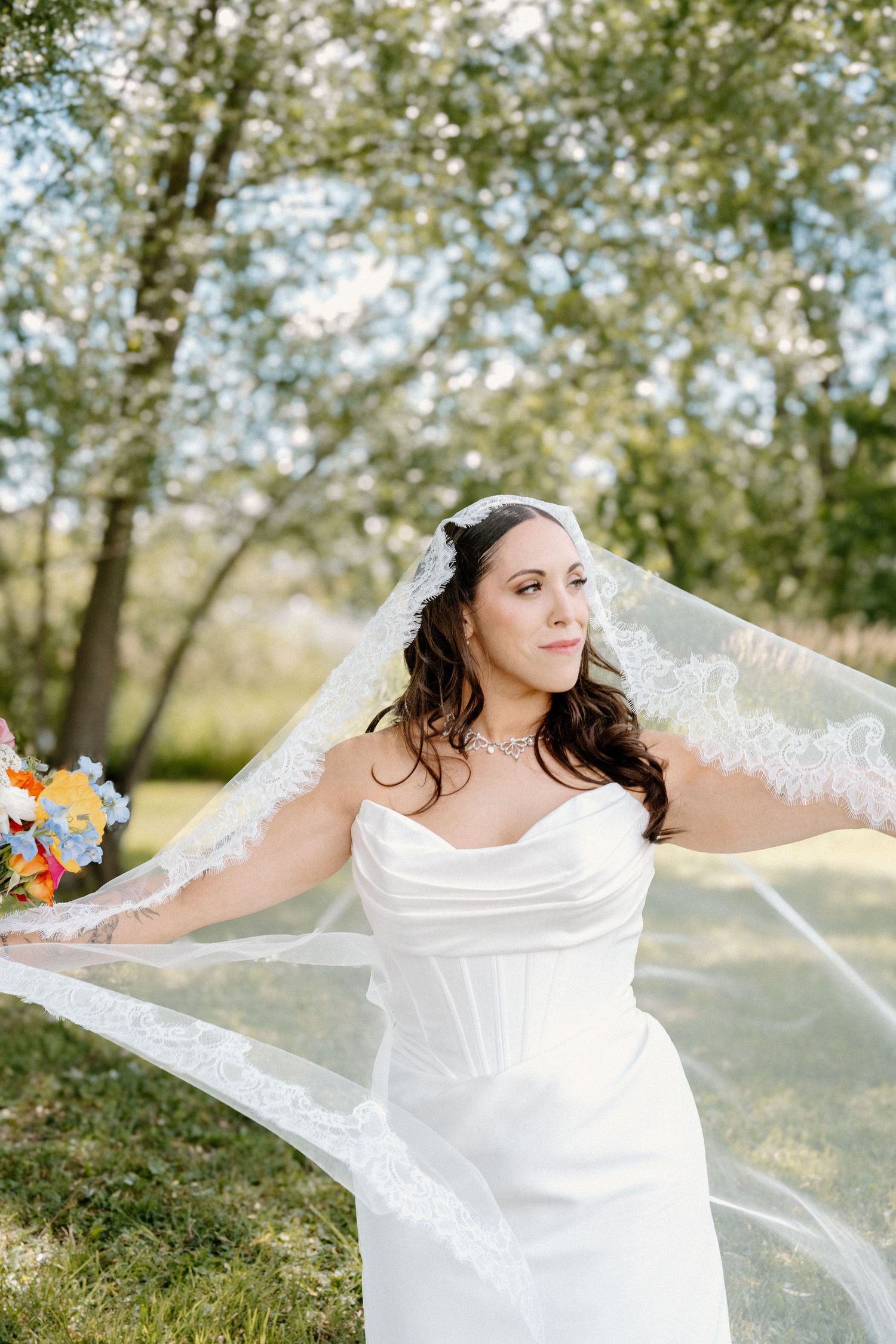 Bride in white strapless gown with flowing lace veil holding colorful bouquet outdoors among green trees.