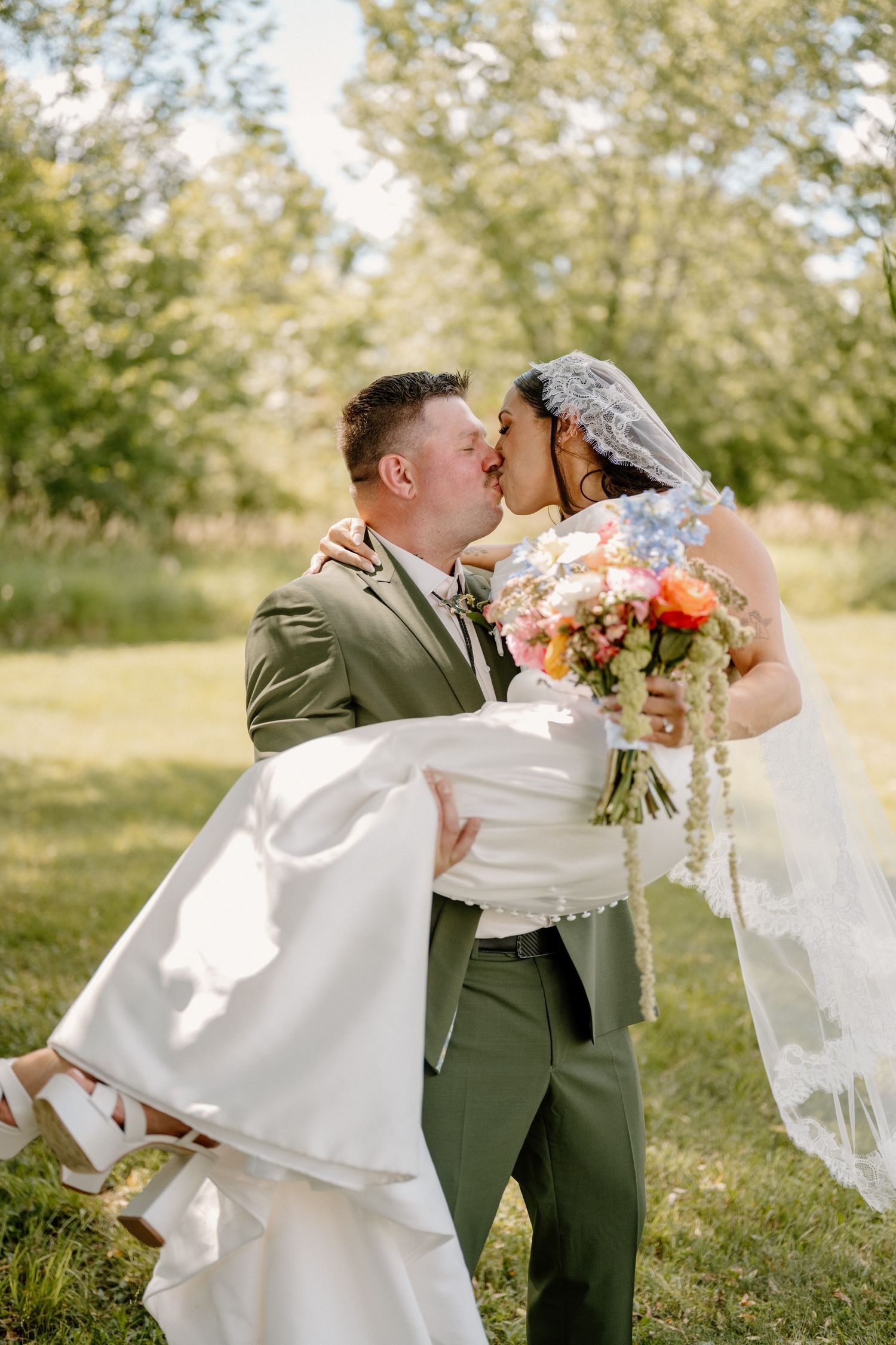 Groom in olive suit carries bride in white gown as they kiss outdoors, bride holding colorful bouquet. Taken in Wallkill, New York