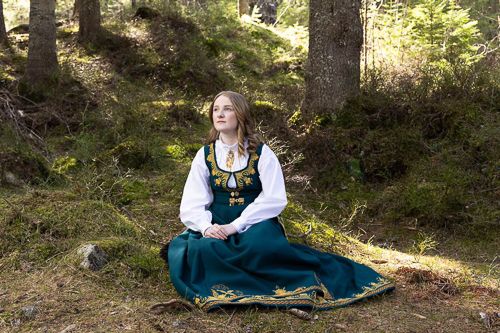 Konfirmant sitter med blå romeriksbunad i skogen. Kofirmasjonsfotografering på Marikollen i Rælingen