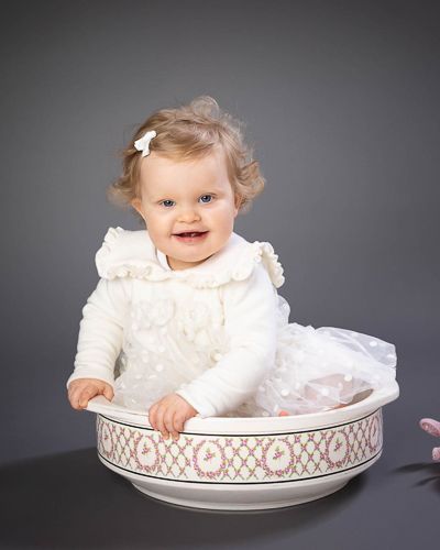 Baby in white dress sits playfully inside decorative china dish against dark studio background.