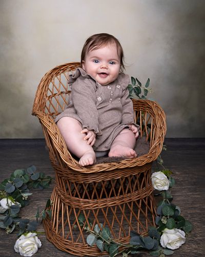 A baby in a gray romper sits in a woven wicker basket with white roses and greenery as studio props.