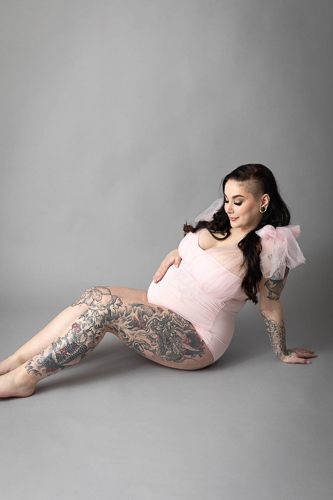 A pregnant woman in a pink dress with elaborate tattoos poses gracefully on a gray studio floor.
