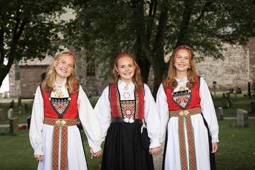 Tre konfirmantjenter i bunad som går hånd i hånd forran en kirke.