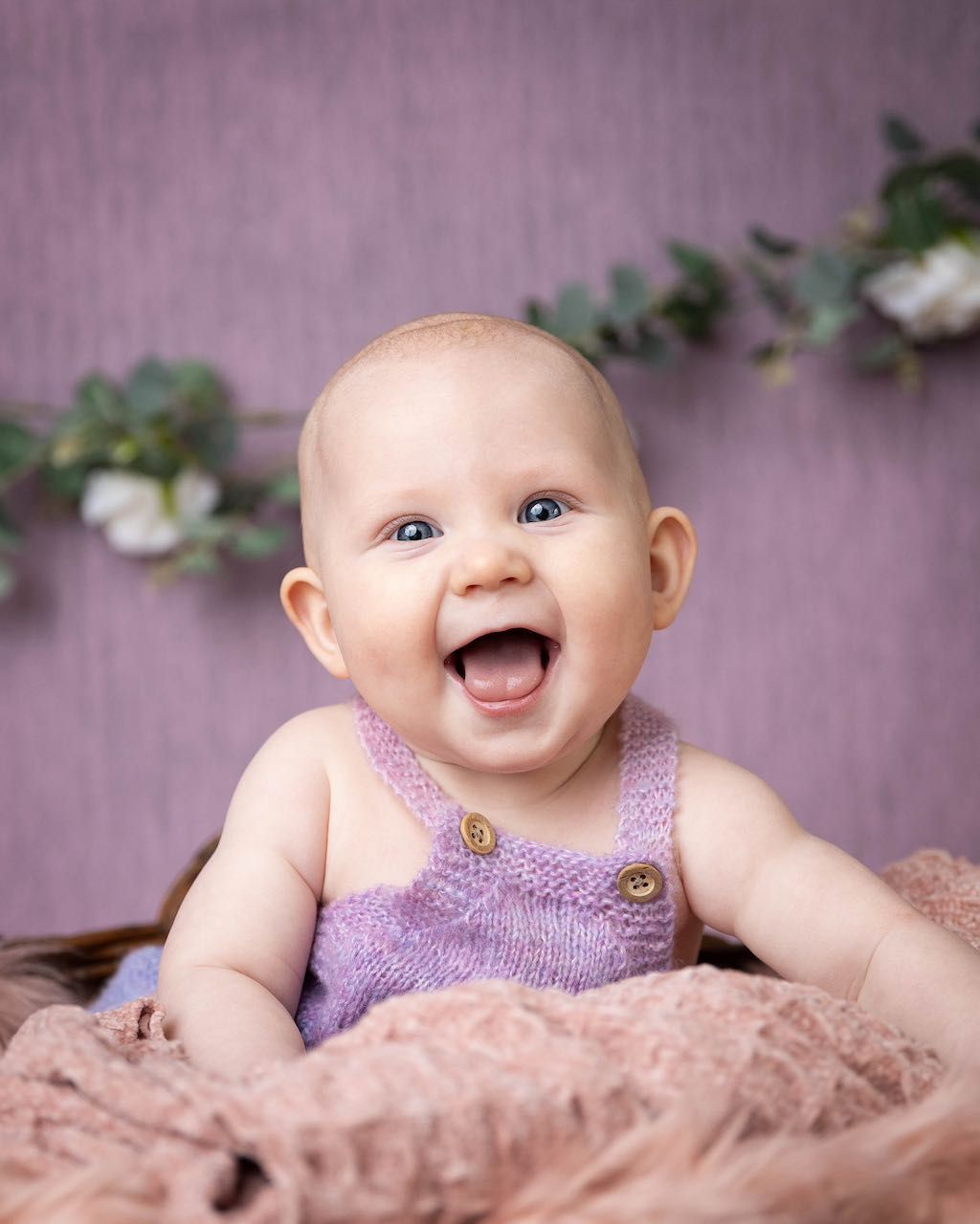 Happy baby wearing purple knit overalls sits among floral decorations against a mauve backdrop.