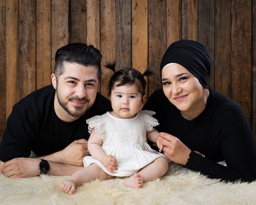 Familiebilde med mamma, pappa og baby på 9 måneder. Foreldrene har svarte klær og baby hvit kjole. Mammaen bærer en hidjab.