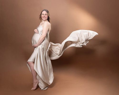 Gravidfoto med beige silkestoff drappert rundt kroppen med brun bakgrunn. Gravidfotografering i studio på Fjerdingby ved Lillestrøm