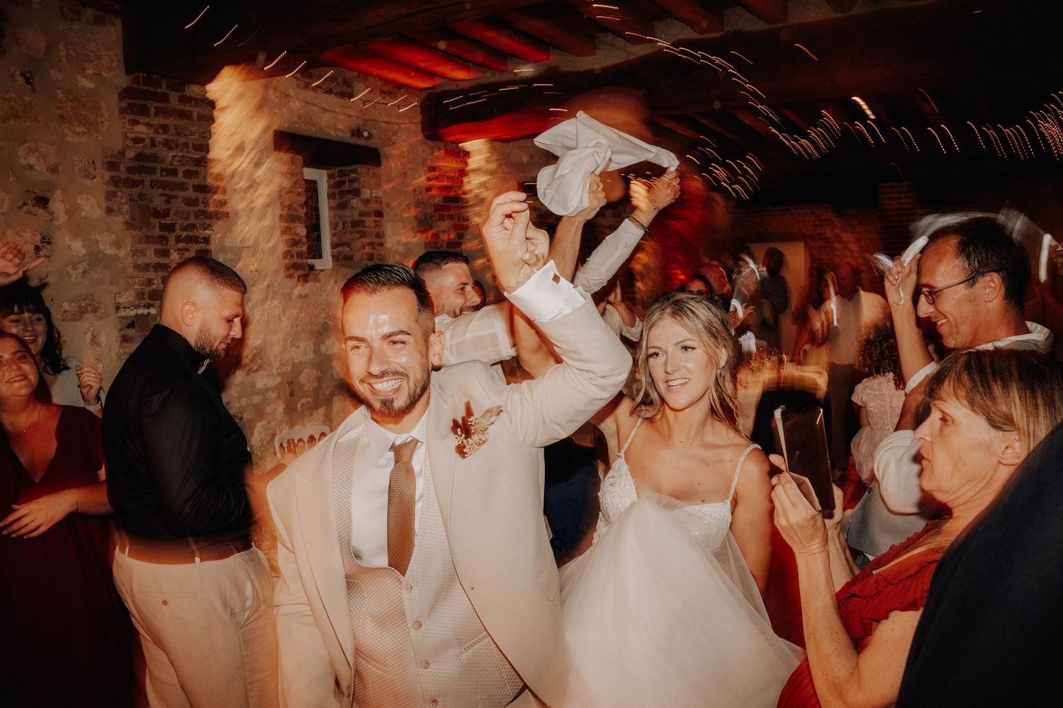 People dancing and celebrating at a wedding reception in a rustic venue with brick walls and warm lighting.