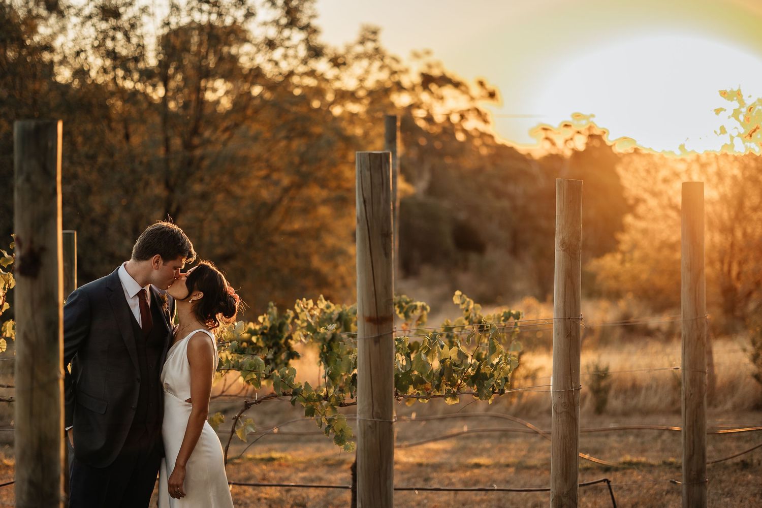 A romantic couple shares an intimate moment at sunset in a rustic outdoor setting with wooden fence posts.