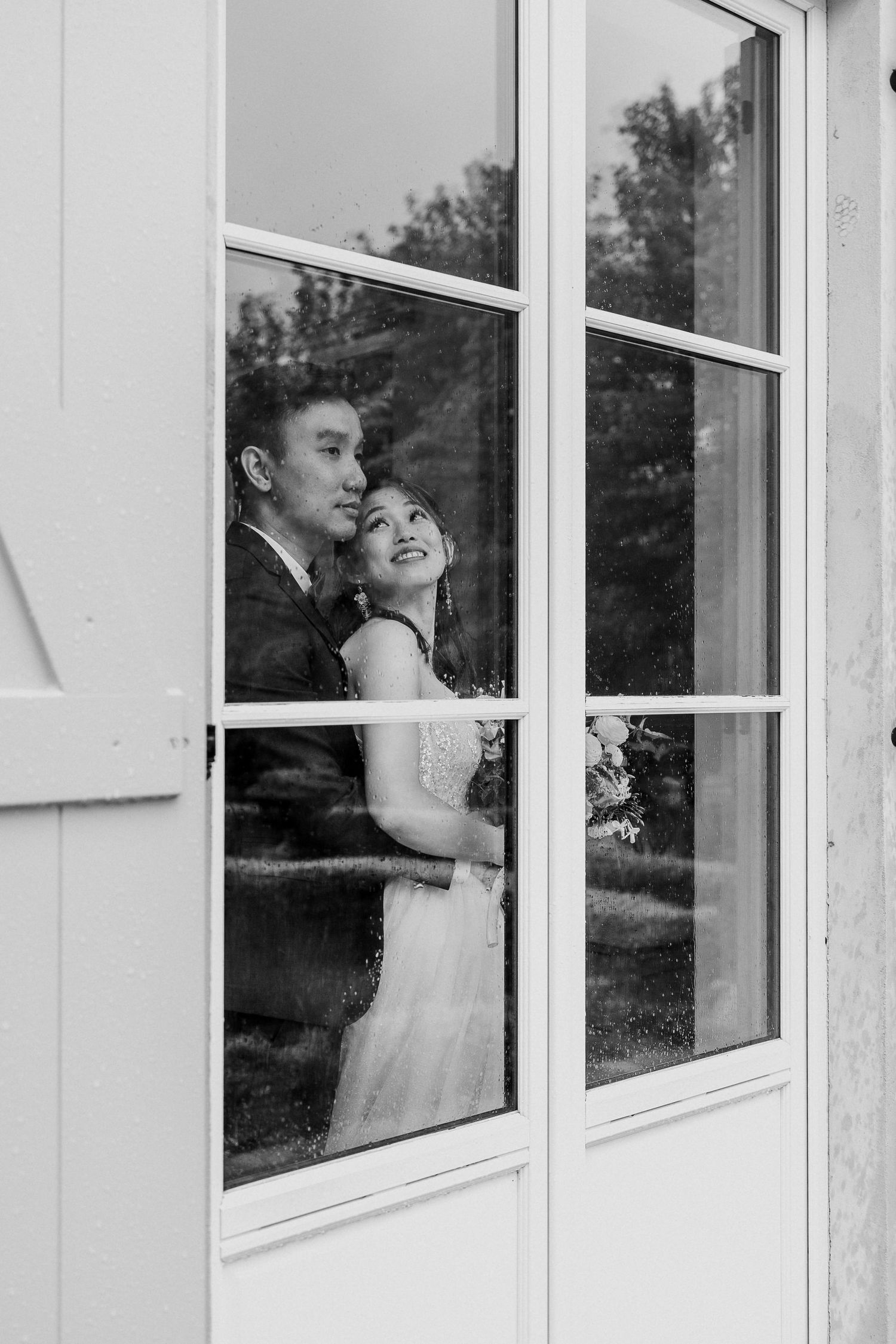 A romantic black and white shot of a couple embracing by a window pane.