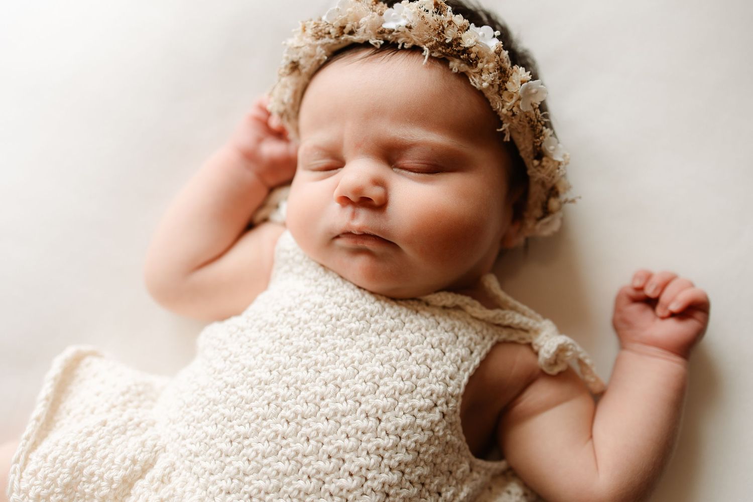 Newborn baby wearing white lace wrap and floral crown sleeps peacefully during professional photo session.