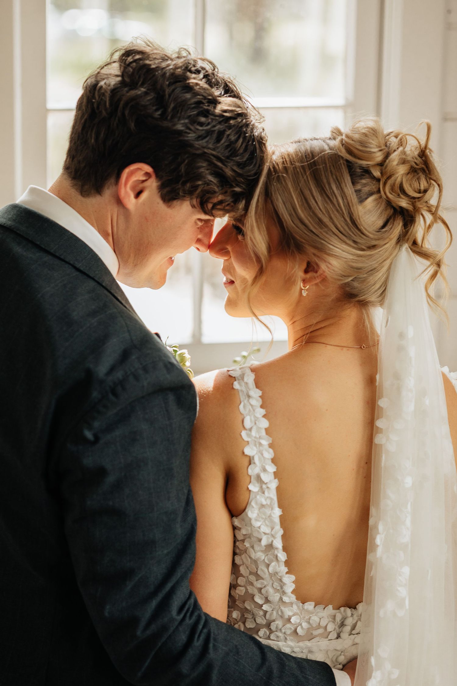 Intimate wedding portrait of couple sharing tender moment by window in natural light.
