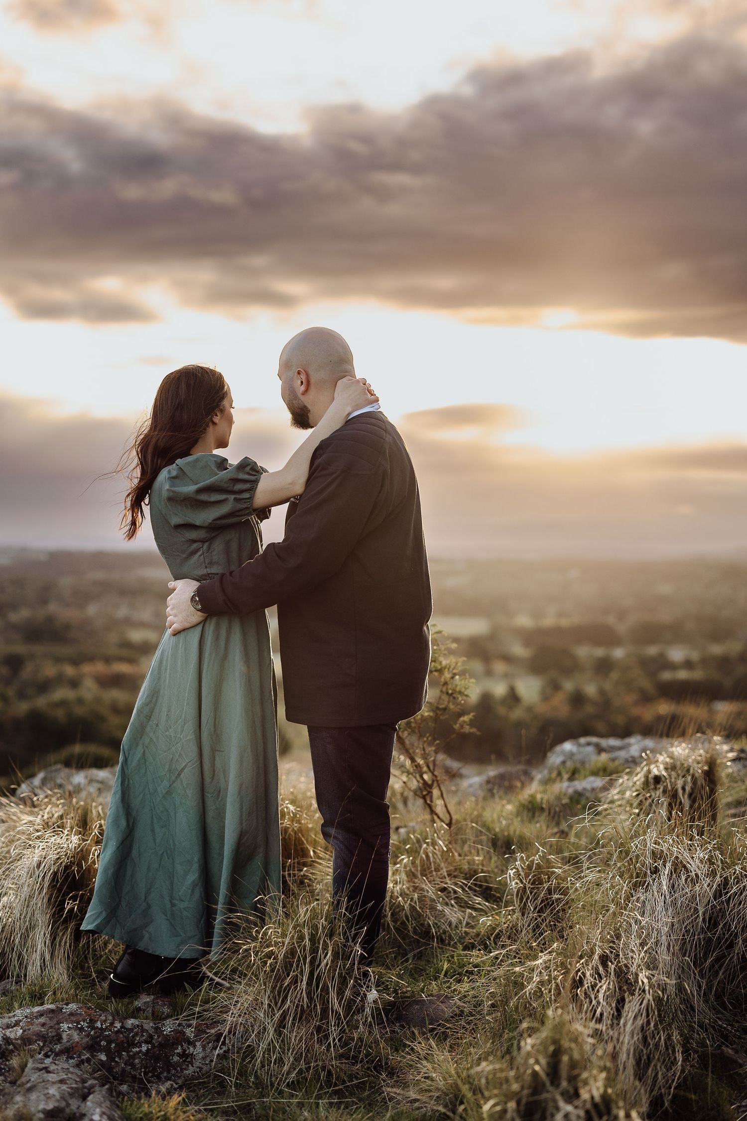 A romantic couple standing on a cliff during sunset in elegant attire.