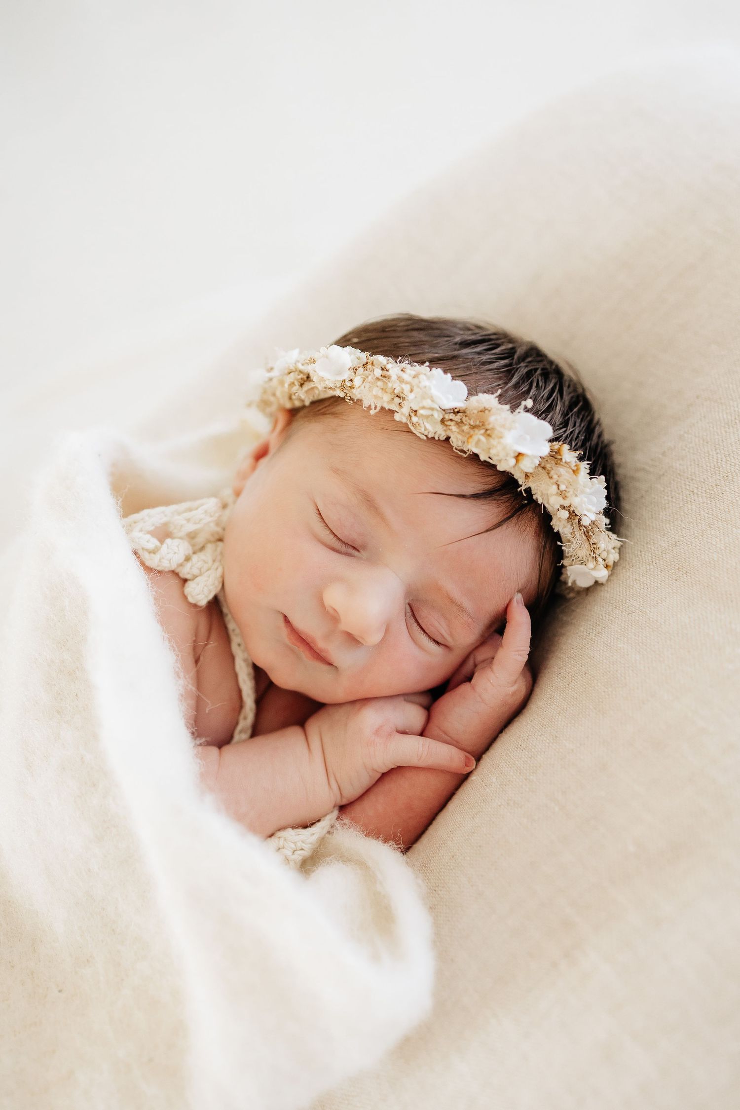 Sleeping newborn baby girl wearing a delicate floral crown, wrapped in a soft cream blanket.