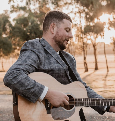 Musician in plaid blazer plays acoustic guitar outdoors during golden hour sunset in wooded area.