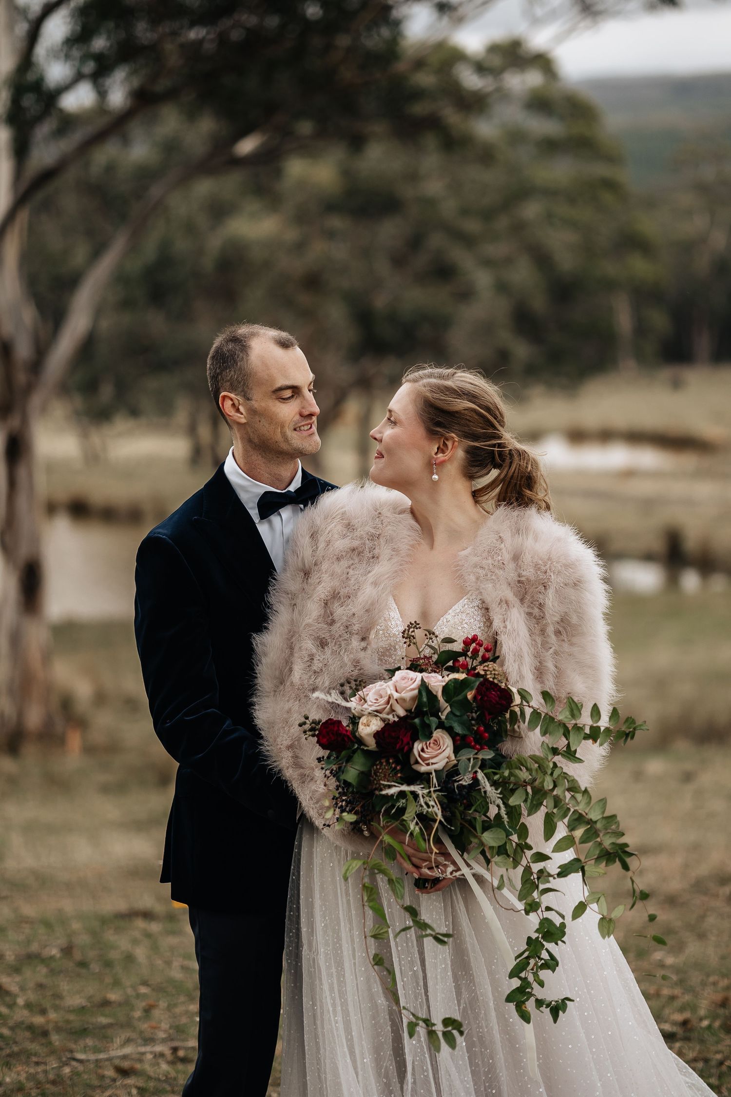 Elegant wedding couple in formal attire pose with bouquet against countryside backdrop.