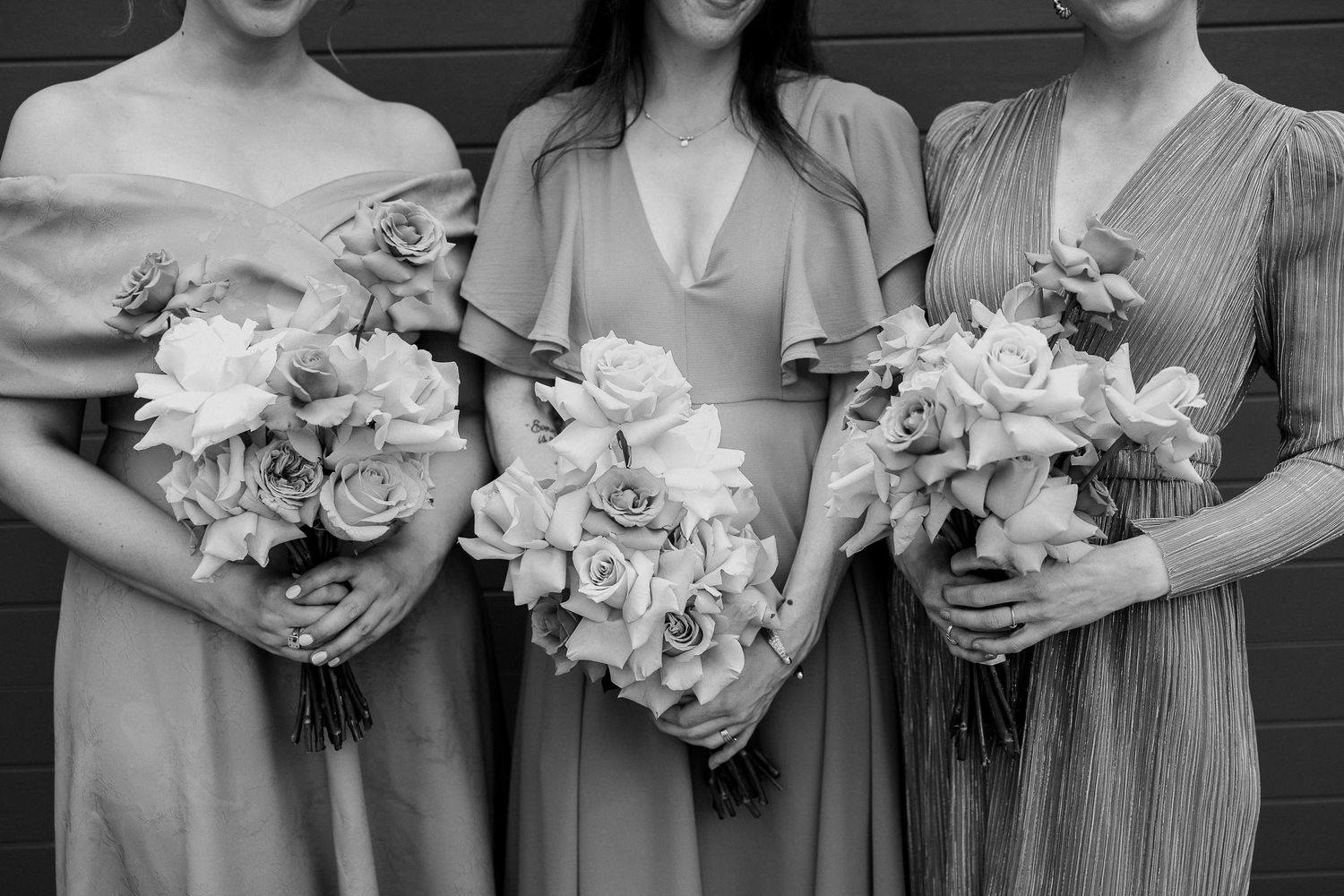 A row of bridesmaids in matching gray dresses hold white floral bouquets against a dark wall.