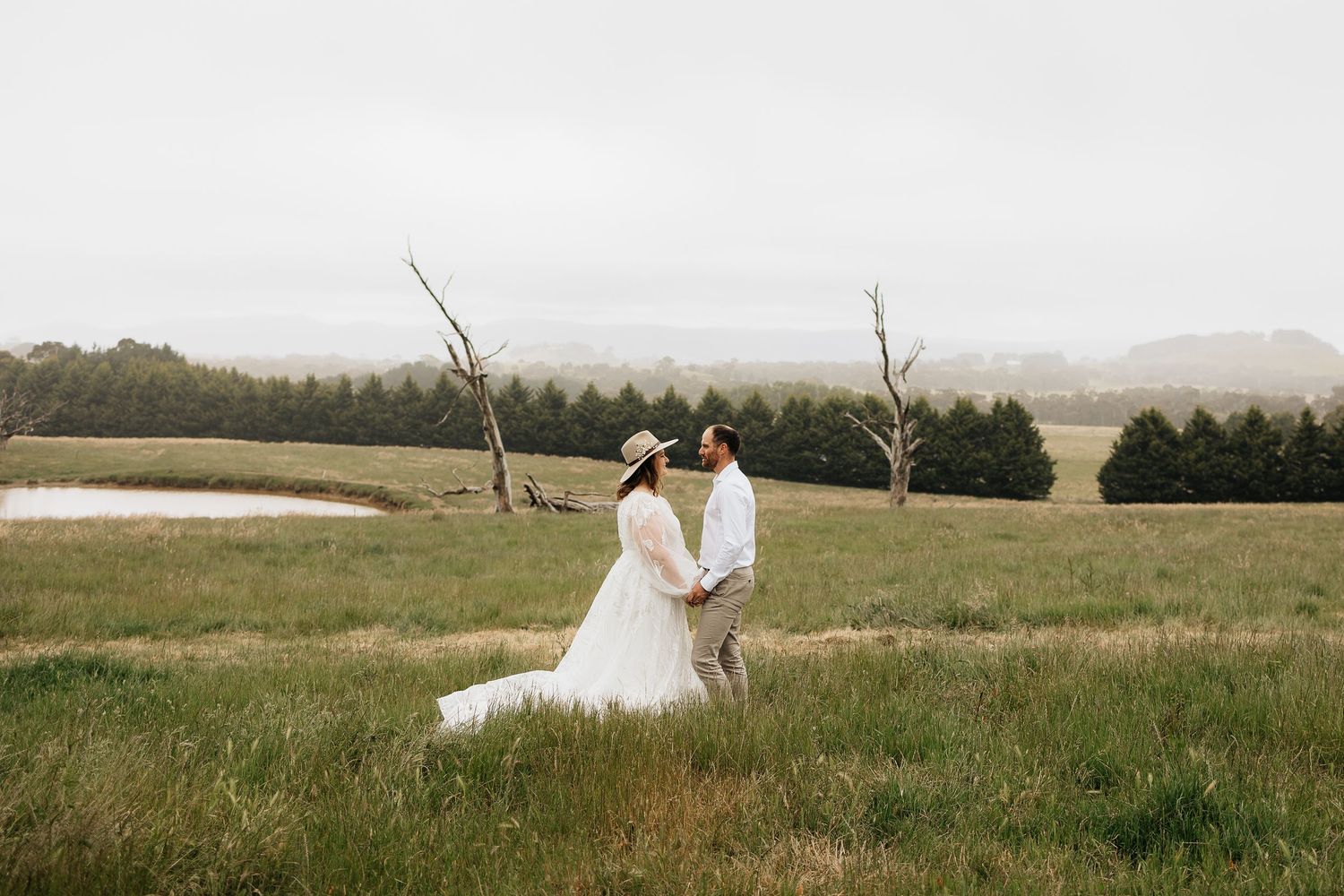 A romantic wedding couple stands together in a vast golden field during sunset with mountains in the distance.