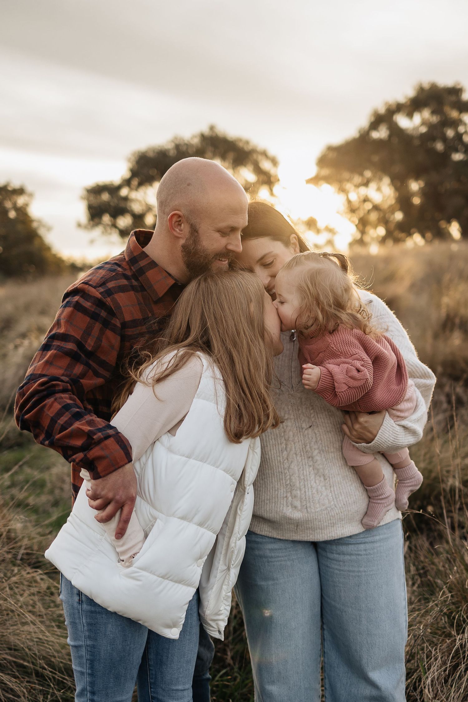 Family sharing warm embrace during golden hour in field with sunset backdrop.