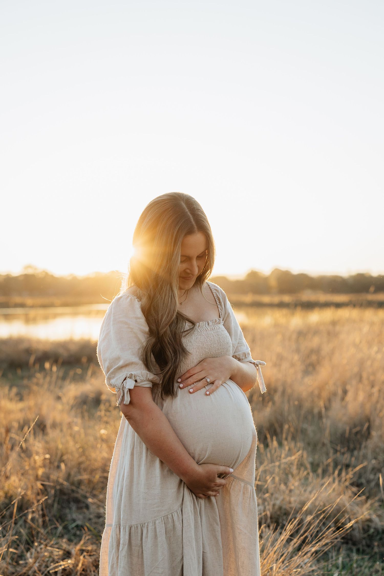 Pregnant woman in beige dress cradling baby bump at golden sunset in a field.