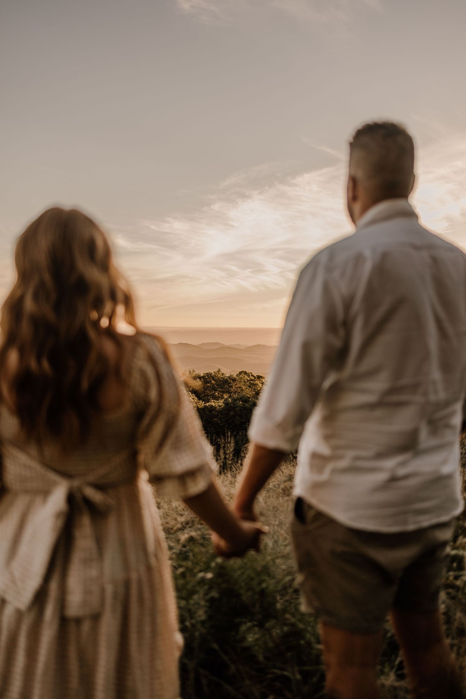 A couple holds hands while watching a scenic sunset over a mountainous landscape in warm golden light.