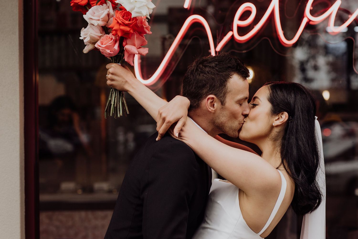 A romantic wedding couple embraces in front of a glowing red neon sign while holding a pink floral bouquet.