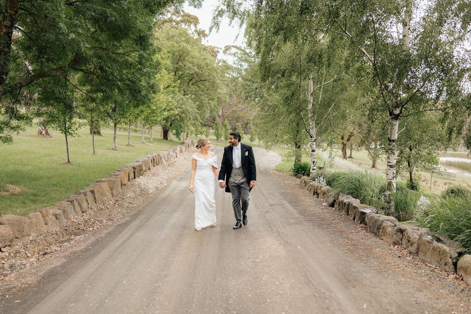 A couple walks together on a tree-lined gravel path bordered by stone walls in a rustic countryside setting.