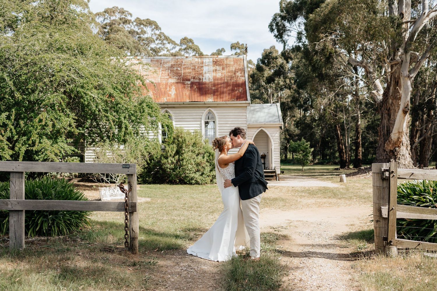 A couple shares an intimate moment on a dirt path between wooden fences with eucalyptus trees in the background.