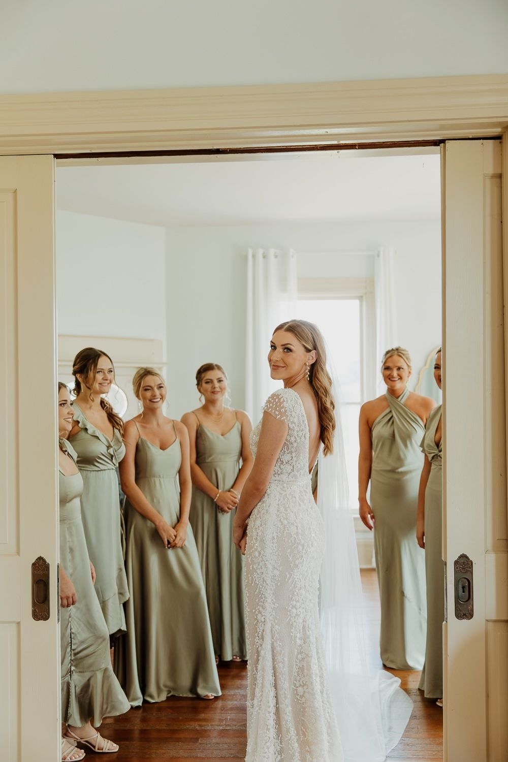 Wedding party wearing sage green bridesmaid dresses gather in doorway with bride in white lace gown.
