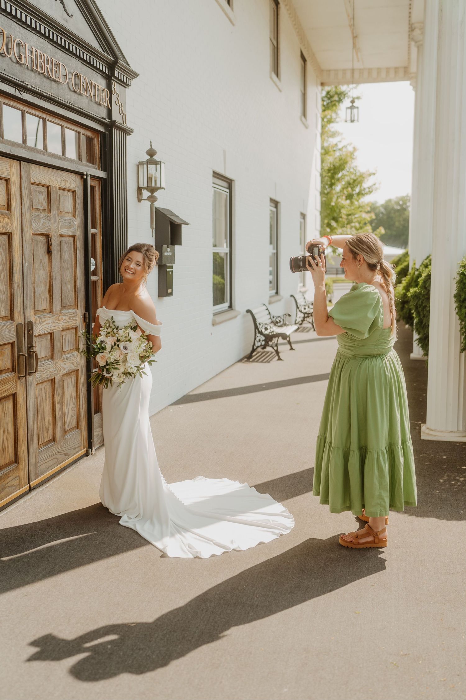 A photographer in a green dress captures wedding photos on a white columned porch with dramatic shadows.