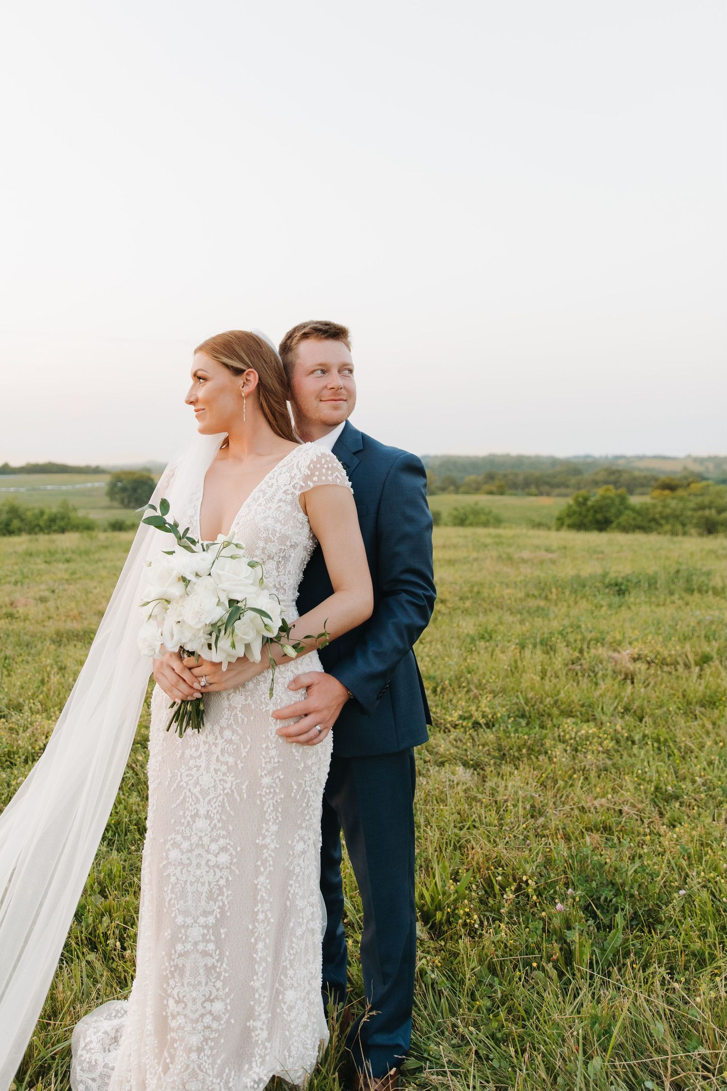 A romantic wedding couple poses in a lush green field at sunset, with the bride holding a white floral bouquet.