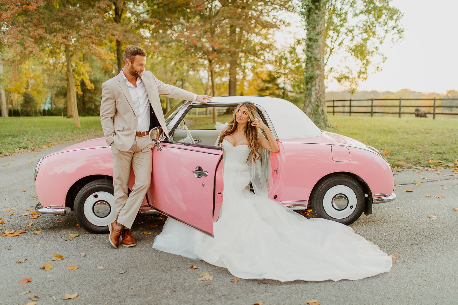 A couple poses with a vintage pink car in an autumn setting during a wedding photo session at sunset.