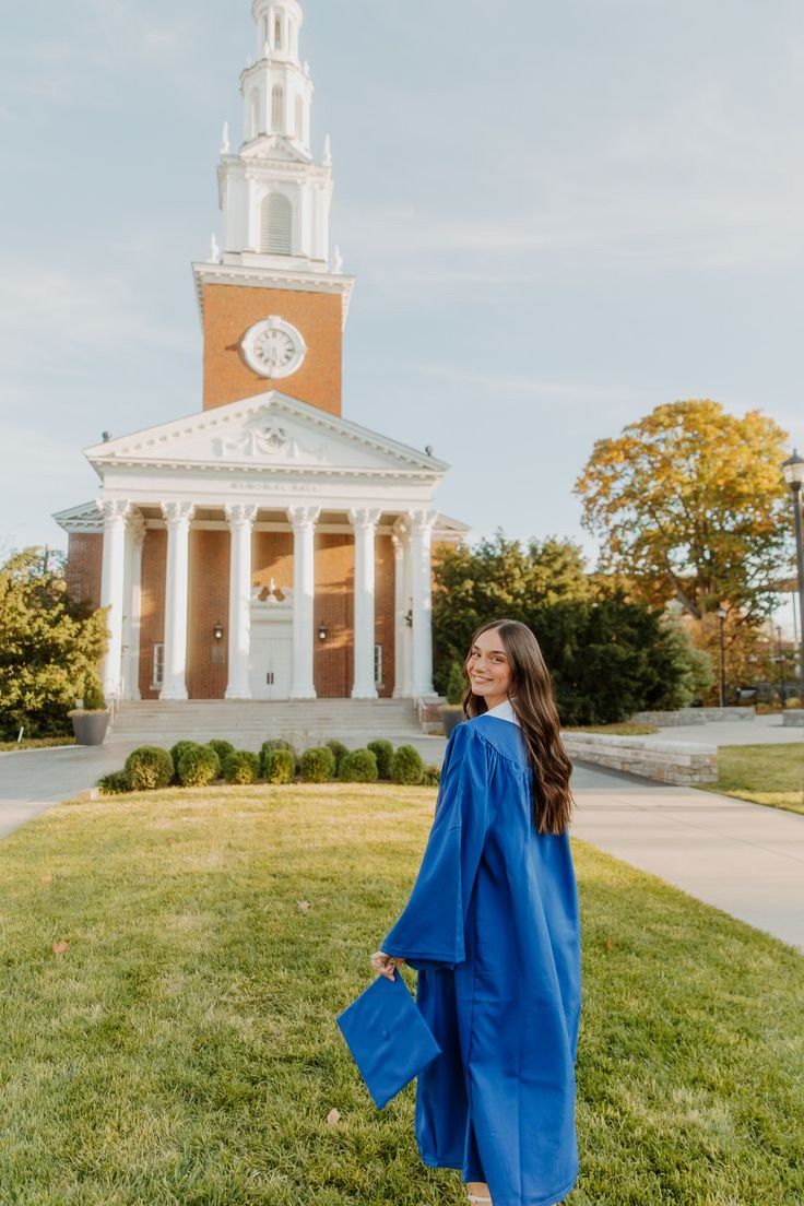 A graduate in blue cap and gown stands on a lawn in front of a grand university chapel with white columns and clock tower.