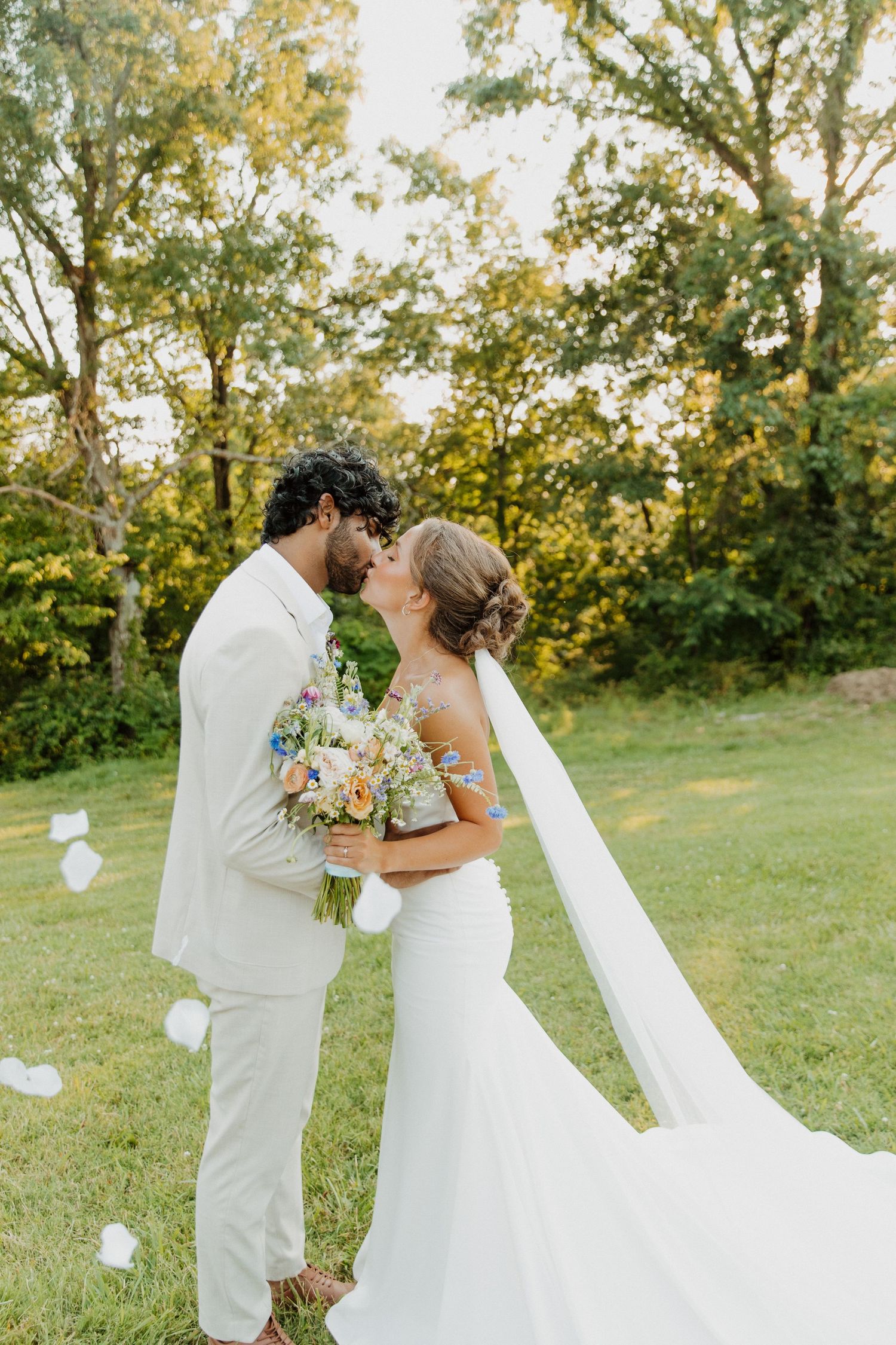 A couple in elegant white wedding attire embrace in a sunlit garden setting with a flowing bridal veil and floral bouquet.