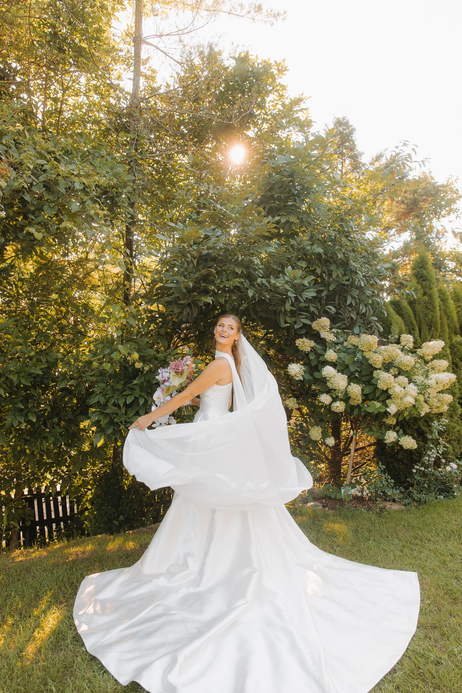 A bride in a flowing white wedding gown twirls in a garden setting with sunlight streaming through trees at golden hour.