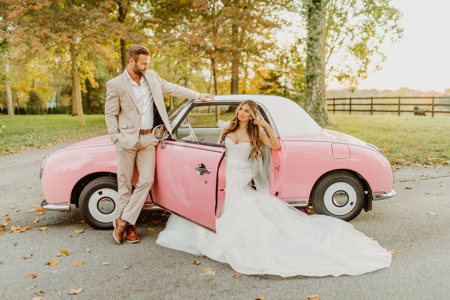 A couple in wedding attire poses together next to a vintage pink car in an autumn setting.