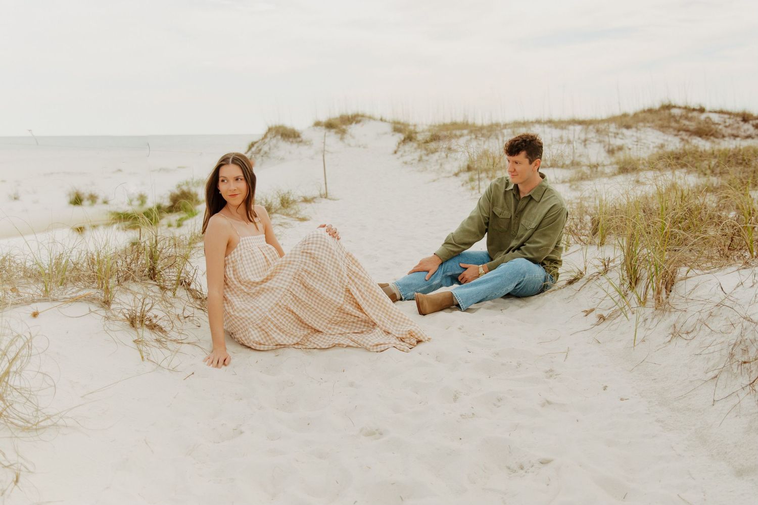 Couple sits together on sandy beach dunes during golden hour in romantic coastal photoshoot.
