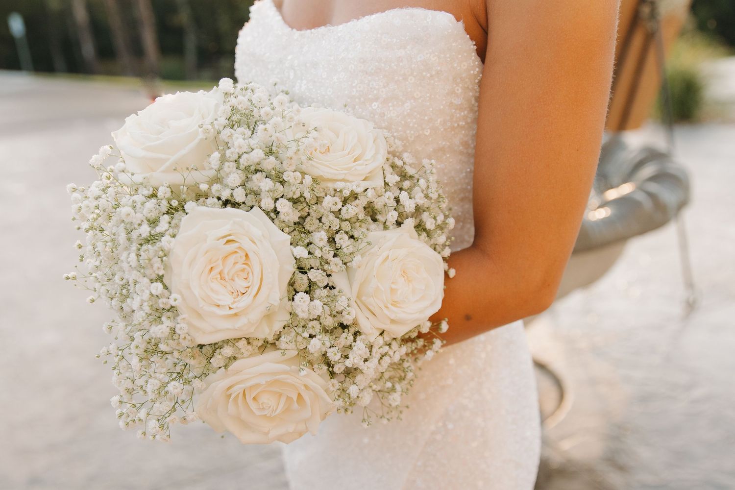 A delicate white rose and baby's breath bouquet is held against a white wedding dress in natural lighting.