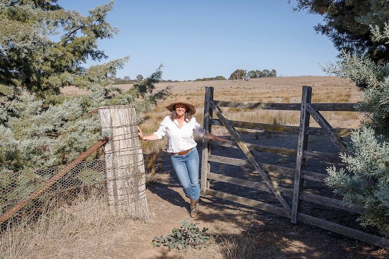 Female farmer with long brown hair in wide brim hat leaning on rustic wooden farm gate. Editorial photography for Graziher magazine.