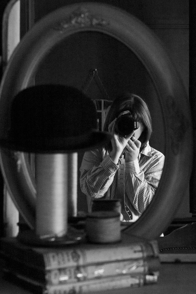 Black and white portrait photo by Em Callaghan Photography, self portrait of a woman in the mirror with a bowler hat & books in foreground.