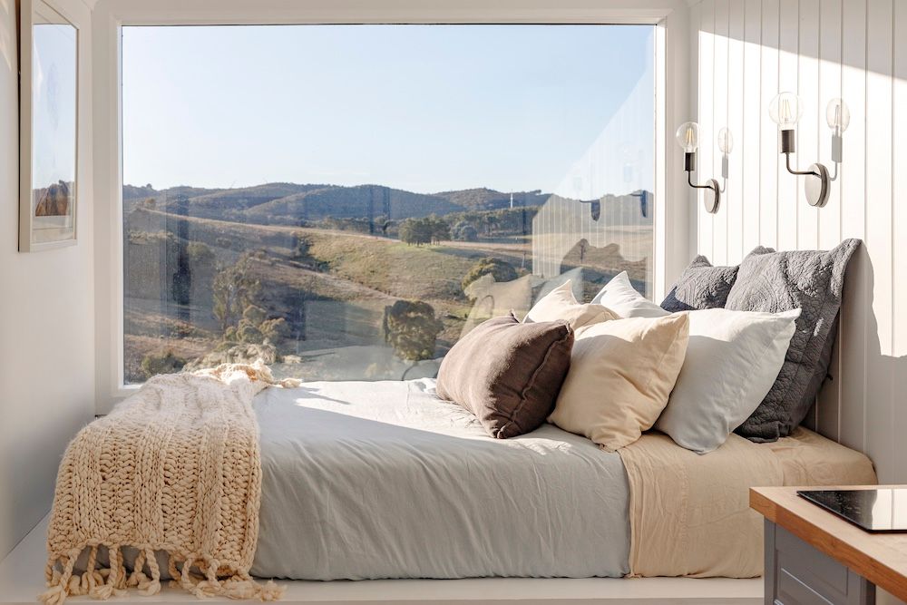 Modern tiny house bedroom with neutral bedding, wall sconce lighting, large window with countryside hills view, interior design.