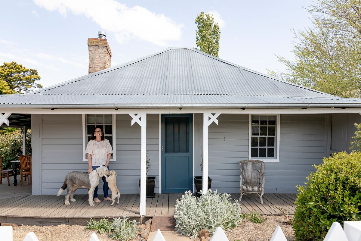 Pale grey blue weatherboard cottage with corrugated iron roof, woman with dog on verandah, Australian country farmhouse exterior