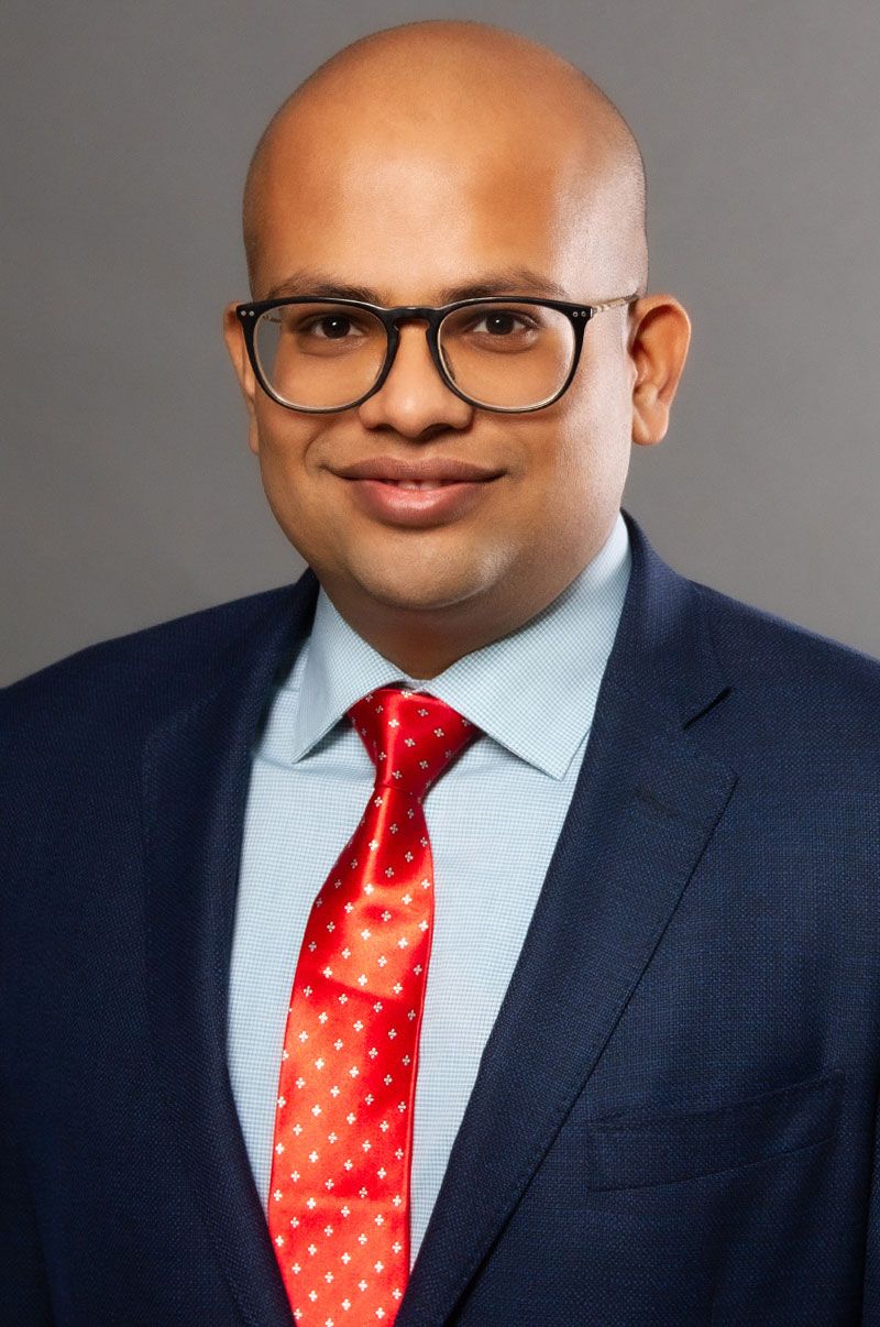 Professional Headshot Suman Koirala wearing dark blue suit jacket blue dress shirt red tie grey background
