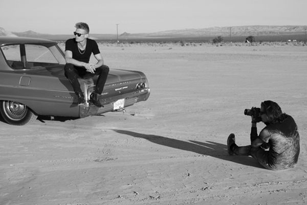 Behind the scenes photo Mark Maryanovich on ground in desert photographing musician Ryan Guldemond sitting on trunk of vintage car