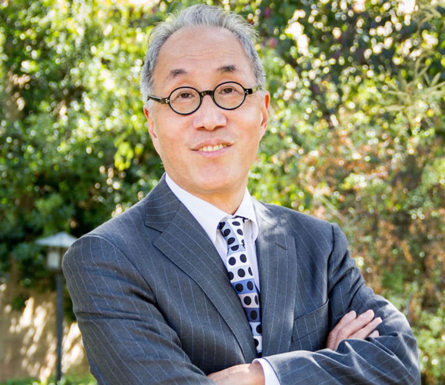 Business headshot featuring Michael Woo hands folded across chest standing in front of trees