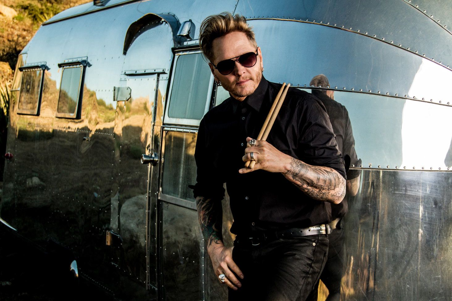 Matt Sorum musician portrait standing against silver airstream trailer holding drumsticks that are leaned on his shoulder