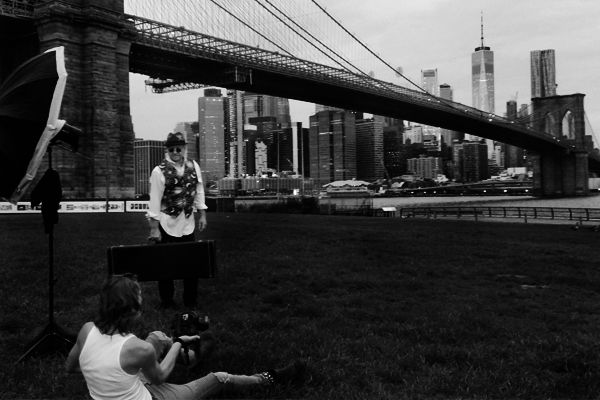 black and white bts image Mark Maryanovich sitting on ground photographing musician Robert Allen standing by Brooklyn Bridge flash directed