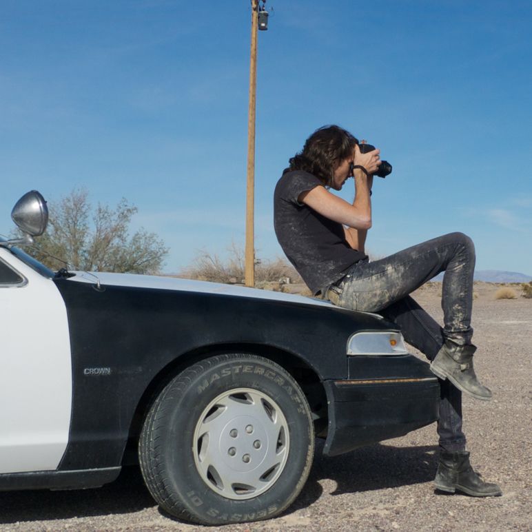 A person with binoculars perches on a police car in a desert setting.