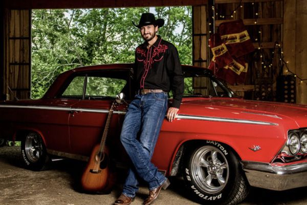 Country music portrait Chance Moore standing next to red classic car guitar leaning on car door beside him parked inside barn