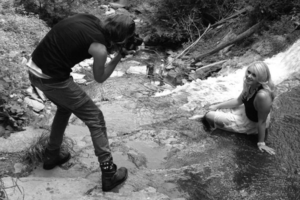 Black and white behind the scenes female musician photoshoot Krista Earle sitting by waterfall while Mark Maryanovich photographs her